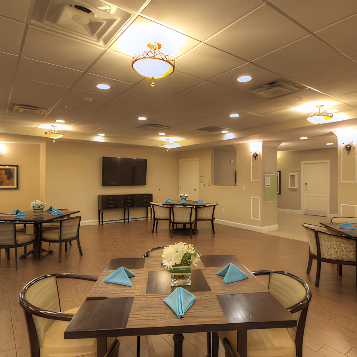 Spacious dining/activity room with multiple tables set with blue folded napkins, chairs, a wall-mounted TV, and warm overhead lighting.