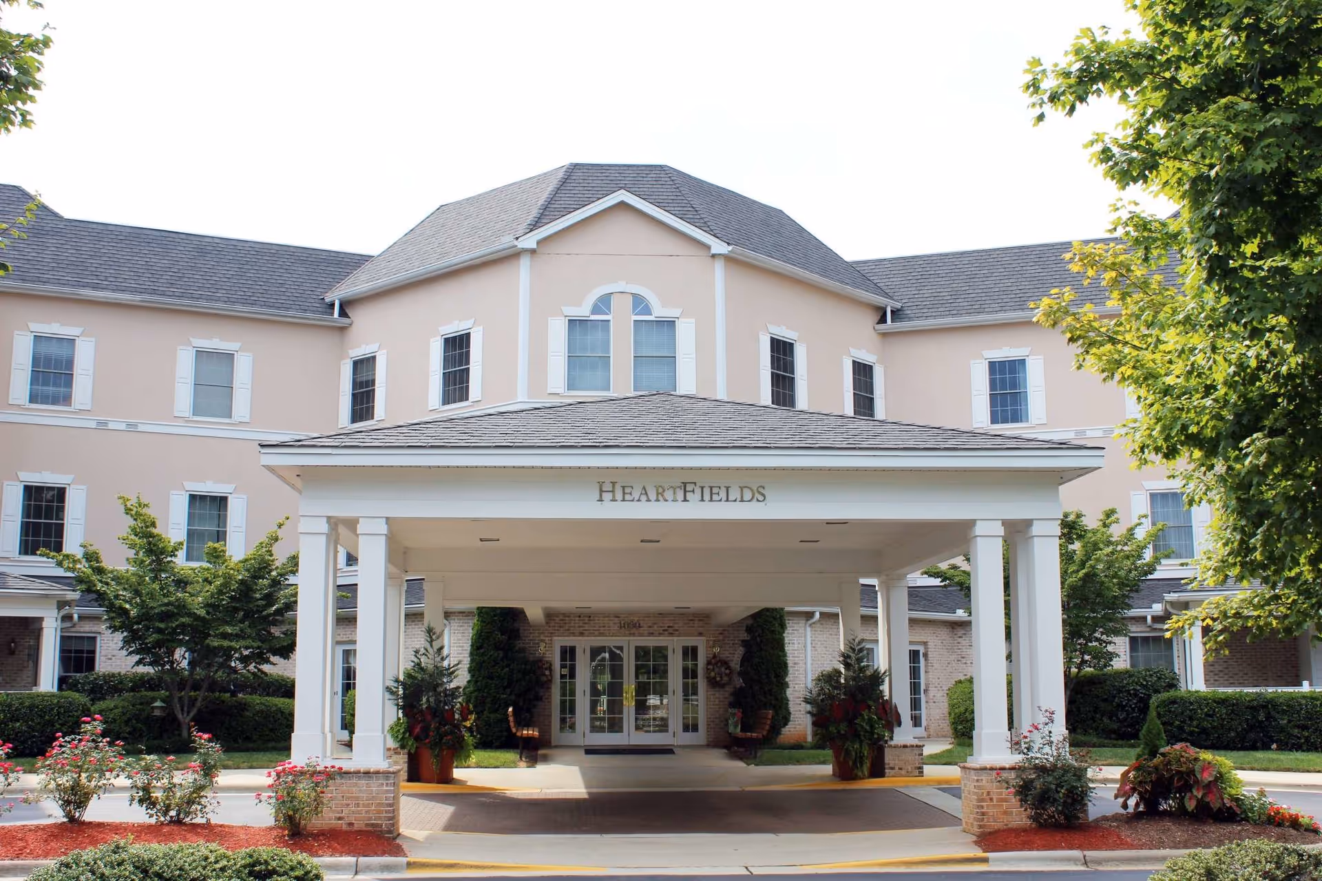Front exterior view of HeartFields at Cary, a multi-story building with beige walls, white trim, and a covered entrance supported by white columns. There are landscaped bushes, trees, and flowers around the entrance area.