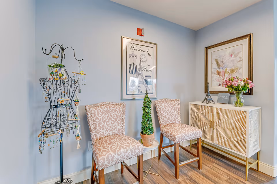 A cozy corner in a senior living facility featuring two patterned upholstered chairs with wooden legs, a small round side table with a potted topiary plant, a decorative wire mannequin adorned with hanging ornaments, and a white cabinet with geometric patterns topped with a small Eiffel Tower model, a vase of pink flowers, and a sign. The walls are painted light blue and decorated with framed artwork.