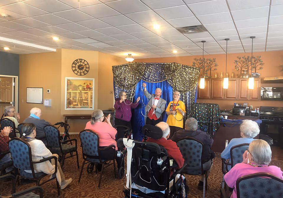 A group of elderly people seated in chairs watching a magic show performed by three entertainers in a senior living facility common area. The performers stand in front of a blue and gold sparkly curtain, holding metal rings for a magic trick. The room has warm-colored walls, a clock, and a kitchen area in the background.