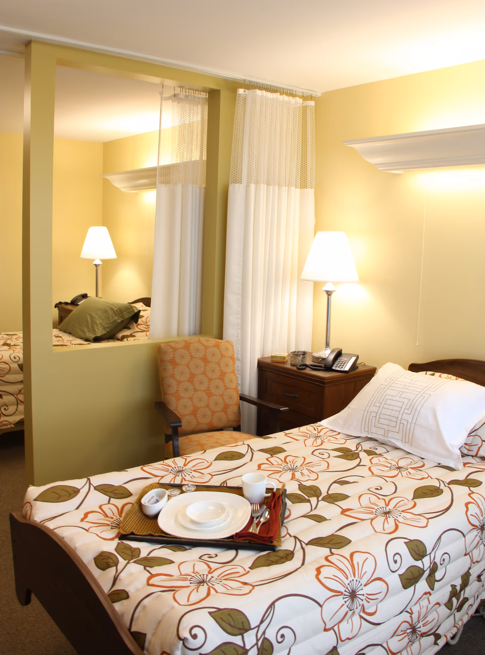 A bright bedroom with a single bed covered in a floral comforter, a tray with dishes on the bed, a bedside table with lamp and phone, and an upholstered chair by a partial room divider and curtain.
