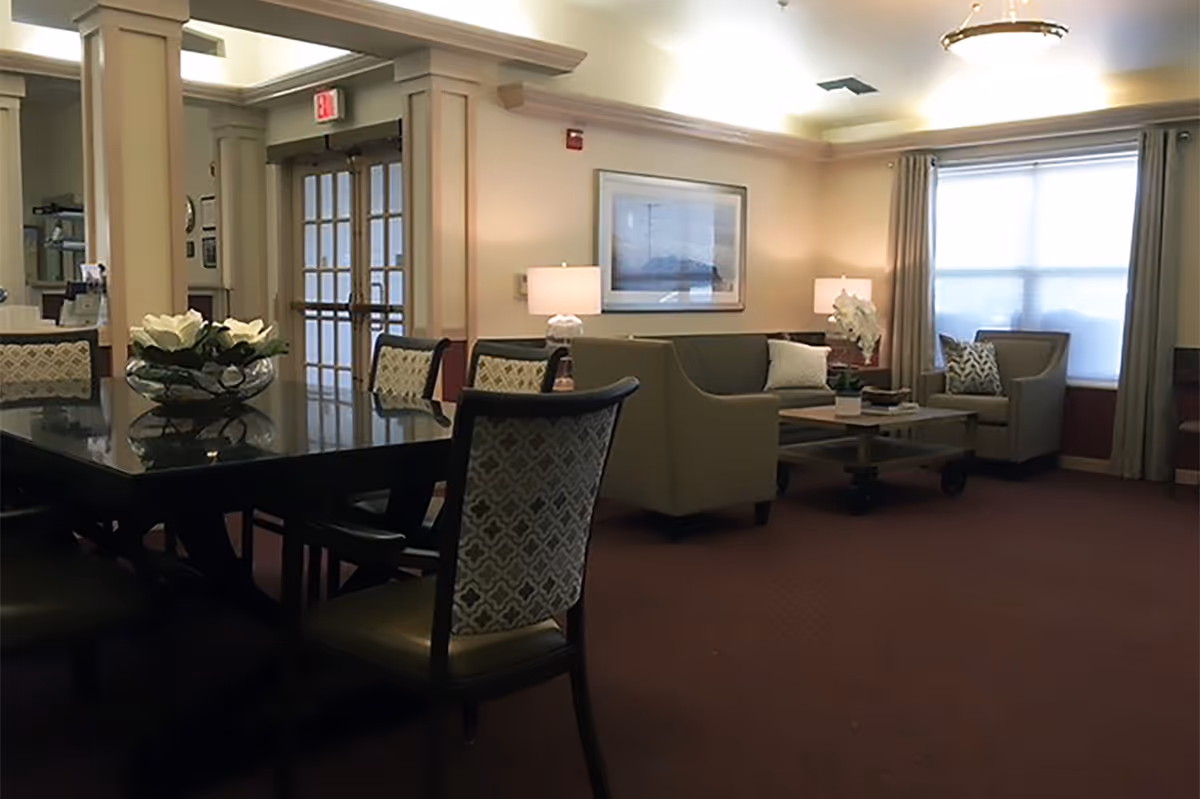 A cozy common area with a dark wooden table and patterned chairs in the foreground, and a seating area with a sofa, armchairs, coffee table, and lamps near a window with curtains in the background. The room has warm lighting and neutral-colored walls.