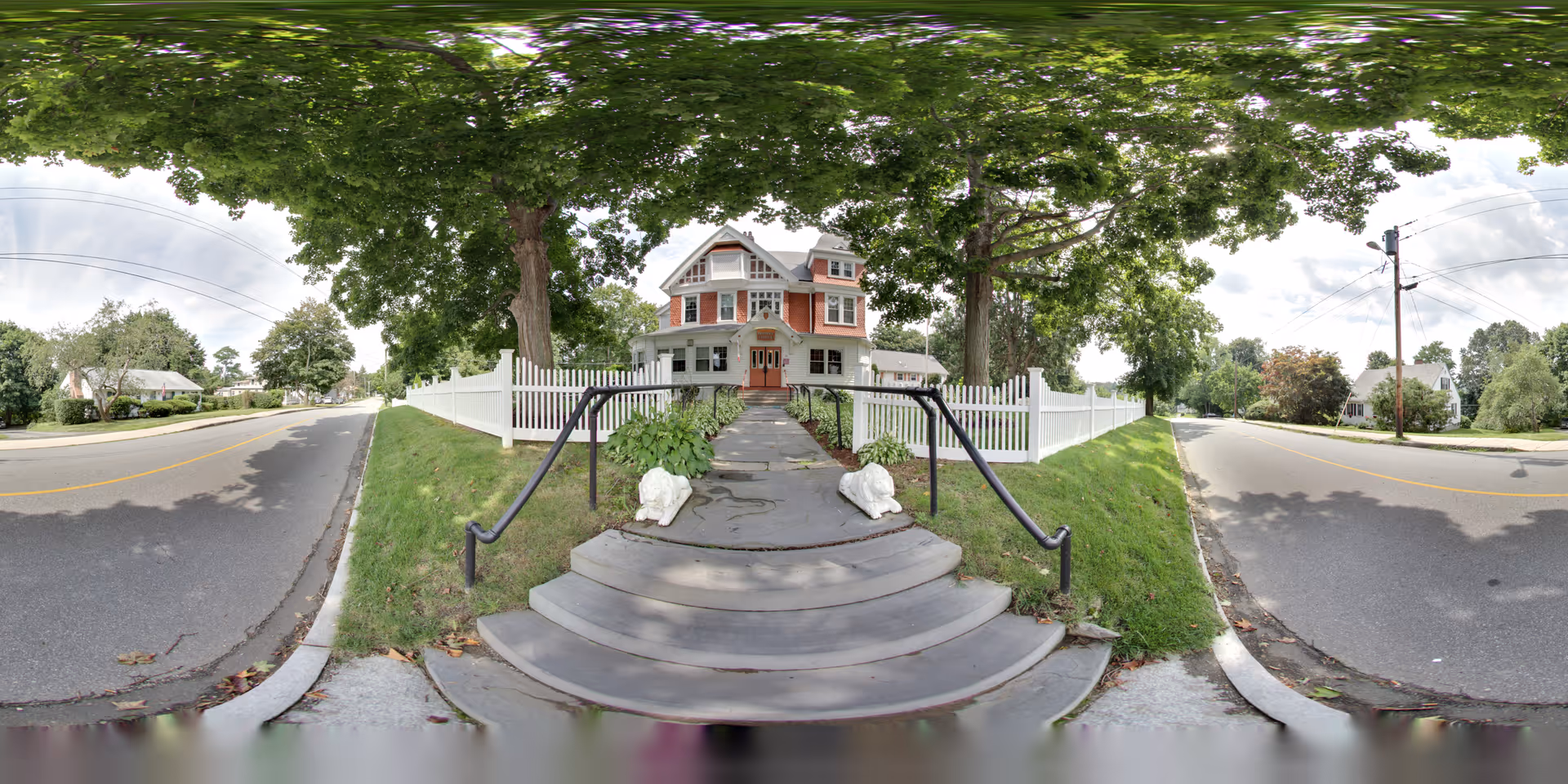 Front exterior view of Riverview Lodge Residential Care Home, a large multi-story house with a white picket fence, surrounded by green trees and situated along a quiet street with sidewalks and power lines.