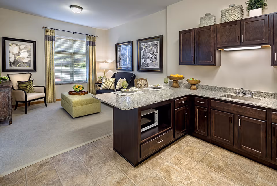 Open-concept interior with a kitchenette featuring dark wood cabinets and a granite island that opens to a cozy living area with seating and a window.