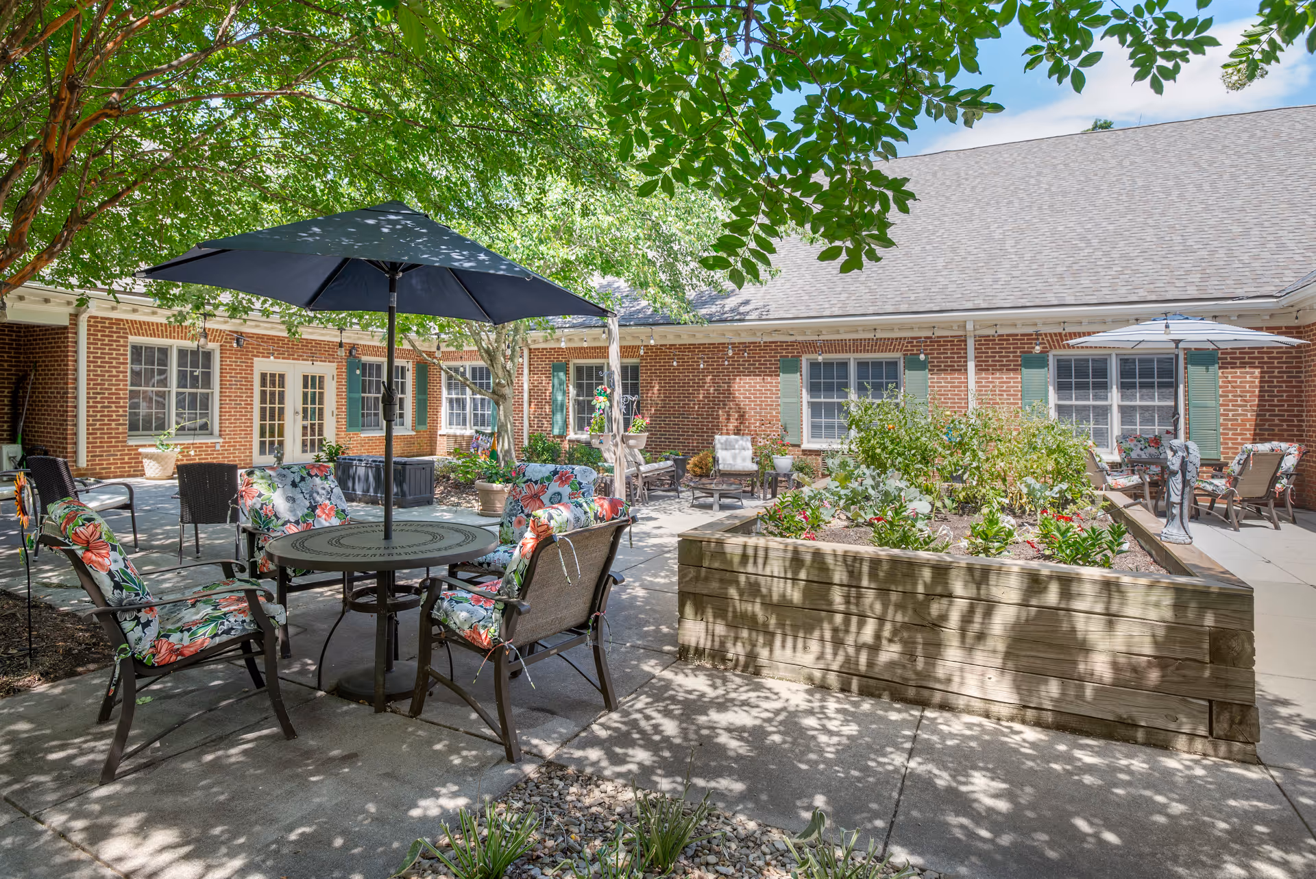 Outdoor courtyard area with patio tables and chairs featuring floral cushions and umbrellas. There is a raised garden bed with plants and flowers, surrounded by a brick building with green shutters and multiple windows. Trees provide shade over the seating area.