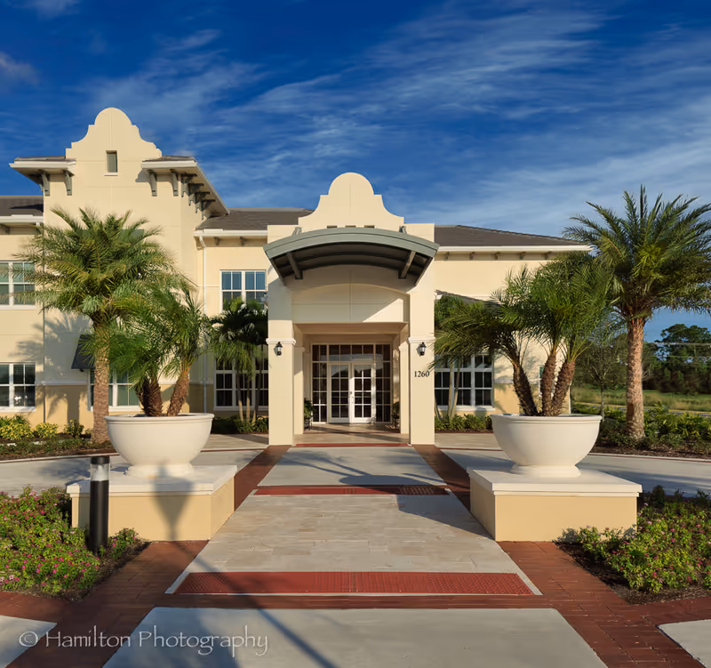 Front exterior view of a light beige building with a covered entrance, large windows, and two large planters with palm trees on either side of the walkway. The sky is clear and blue.