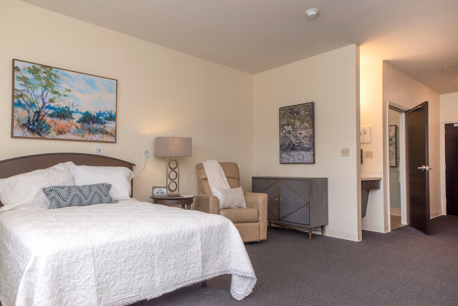 A furnished bedroom with a made bed, bedside table and lamp, armchair, dresser, and wall artwork.