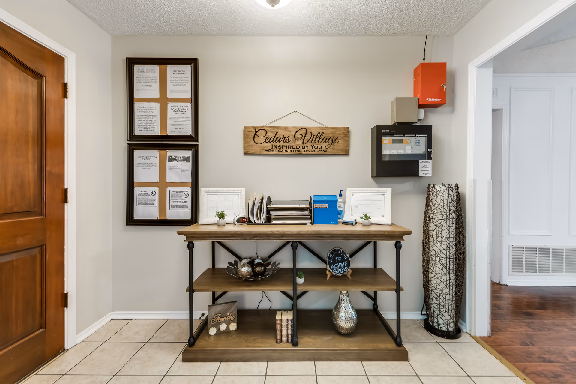 An interior hallway area with a wooden door on the left and a wooden shelf against a beige wall. The shelf holds decorative items including small plants, books, and a silver vase. Above the shelf is a wooden sign that reads 'Cedars Village Inspired By You Carrollton Texas'. There are framed notices on the wall to the left and a fire alarm panel and red fire alarm box on the right wall. The floor is tiled and there is an open doorway leading to a room with wooden flooring on the right.