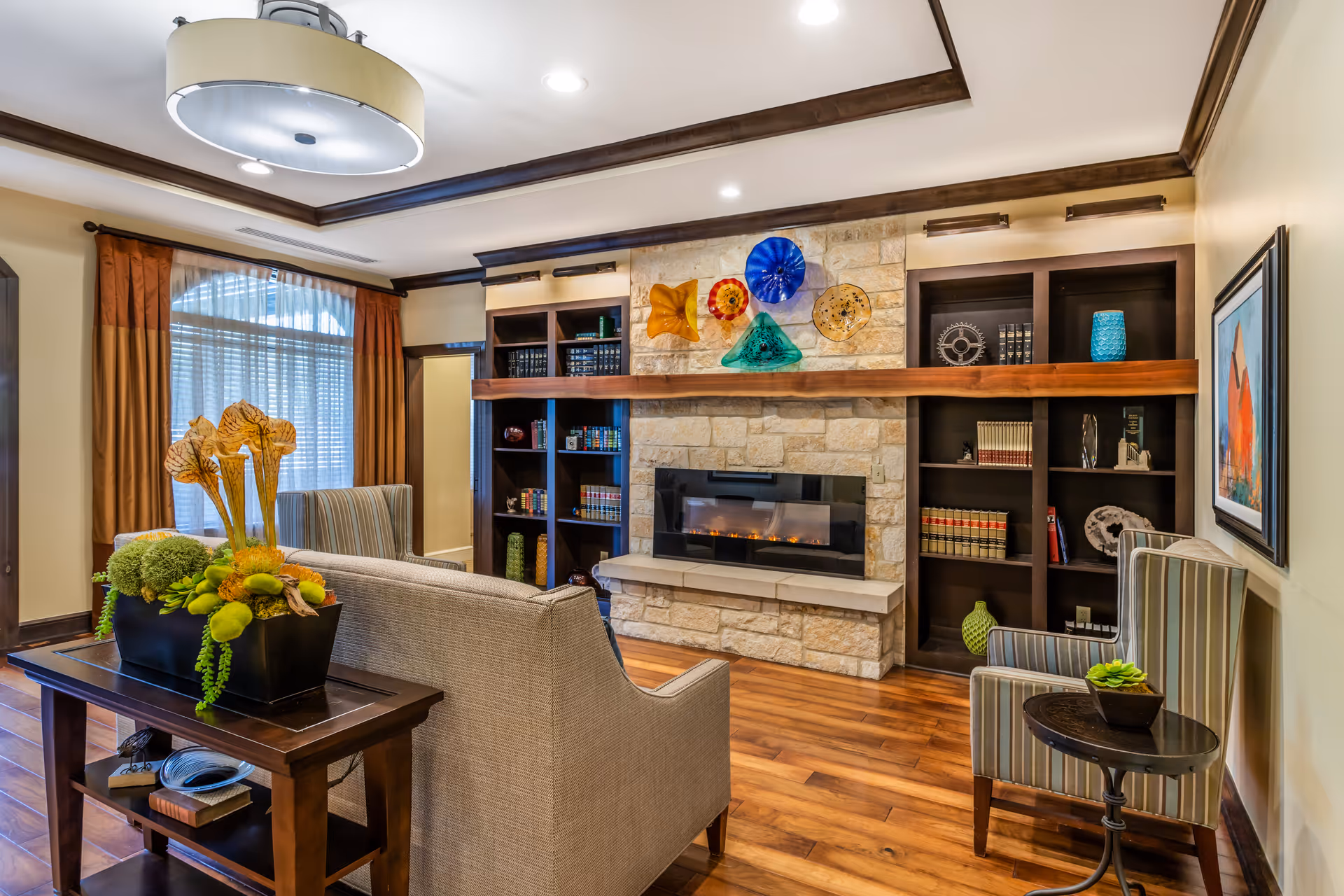 A cozy living room with a stone fireplace featuring colorful glass art above the mantle. The room has wooden floors, built-in dark wood bookshelves filled with books and decorative items, a beige sofa, a striped armchair with a small round side table holding a small plant, and a wooden console table with a floral arrangement. There is a large window with sheer curtains and brown drapes, and a modern ceiling light fixture.