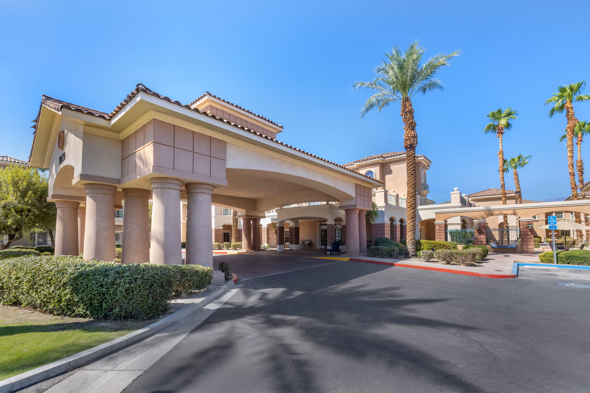 Exterior view of Brookdale Mirage Inn showing the entrance with large columns, a covered driveway, palm trees, and clear blue sky.