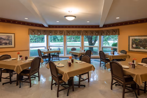 A dining room with several tables covered with beige tablecloths, each set with plates, cups, and small flower vases. The room has large windows with a view of trees and greenery outside. The walls are painted a warm yellow with framed artwork and a decorative valance above the windows.