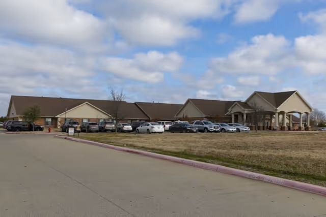 Exterior view of a single-story healthcare and rehabilitation facility building with a large parking lot filled with cars in front. The building has a beige and brick facade with a covered entrance supported by columns. The sky is partly cloudy.