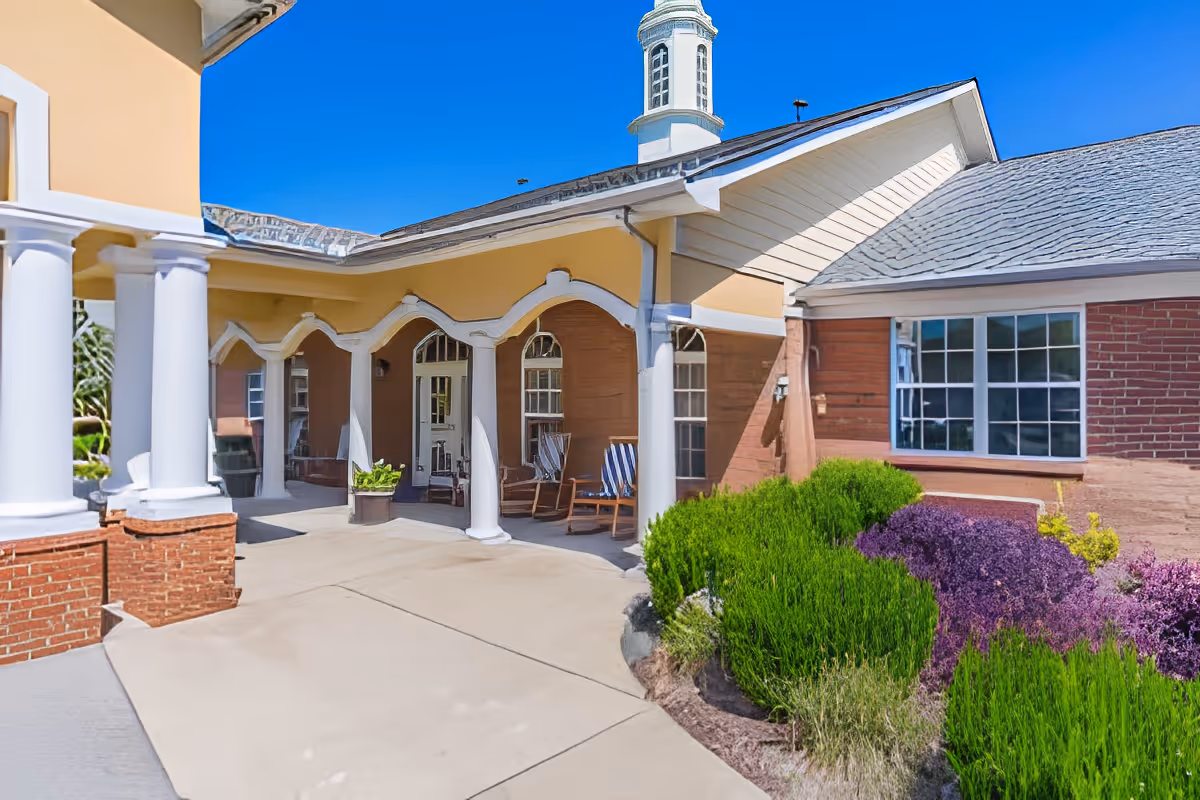 Exterior view of a senior living facility with a covered porch supported by white columns, rocking chairs, and well-maintained landscaping including green and purple bushes under a clear blue sky.