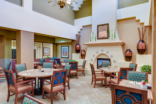 A bright and spacious dining area with multiple wooden tables and chairs featuring blue and green patterned upholstery. The room has a high ceiling with a chandelier, a decorative fireplace with vases and artwork above it, and large windows allowing natural light to enter.