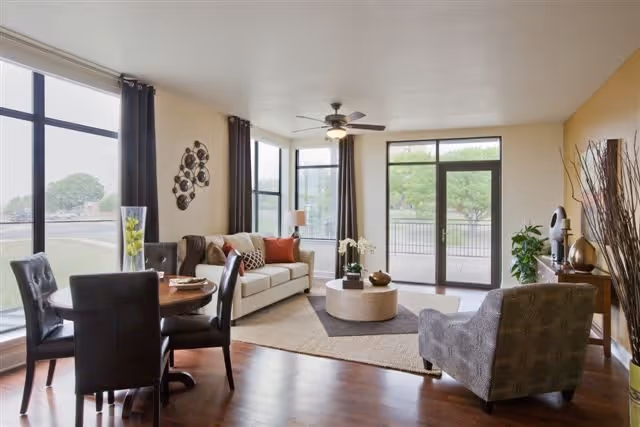 A bright and spacious living room with large windows and glass doors letting in natural light. The room features a beige sofa with colorful cushions, a round coffee table, a patterned armchair, a wooden dining table with four black chairs, and a ceiling fan with a light. The walls are painted in neutral tones with one accent wall in yellow, and there are decorative items including plants, a wall sculpture, and a lamp.