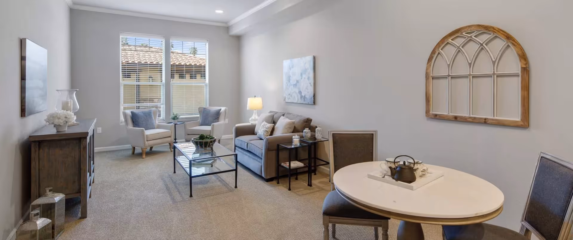 A bright and cozy living room with a gray sofa, two white armchairs with blue cushions, a glass coffee table, and a small round dining table with two chairs. The room has beige carpeting, light gray walls, a large window with blinds, and decorative wall art including a floral painting and a wooden arched window frame.