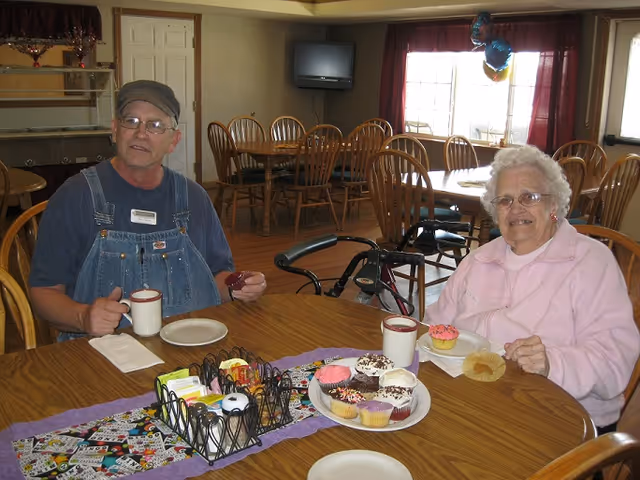 Two older adults sit at a dining table with cupcakes and mugs in a communal dining room.