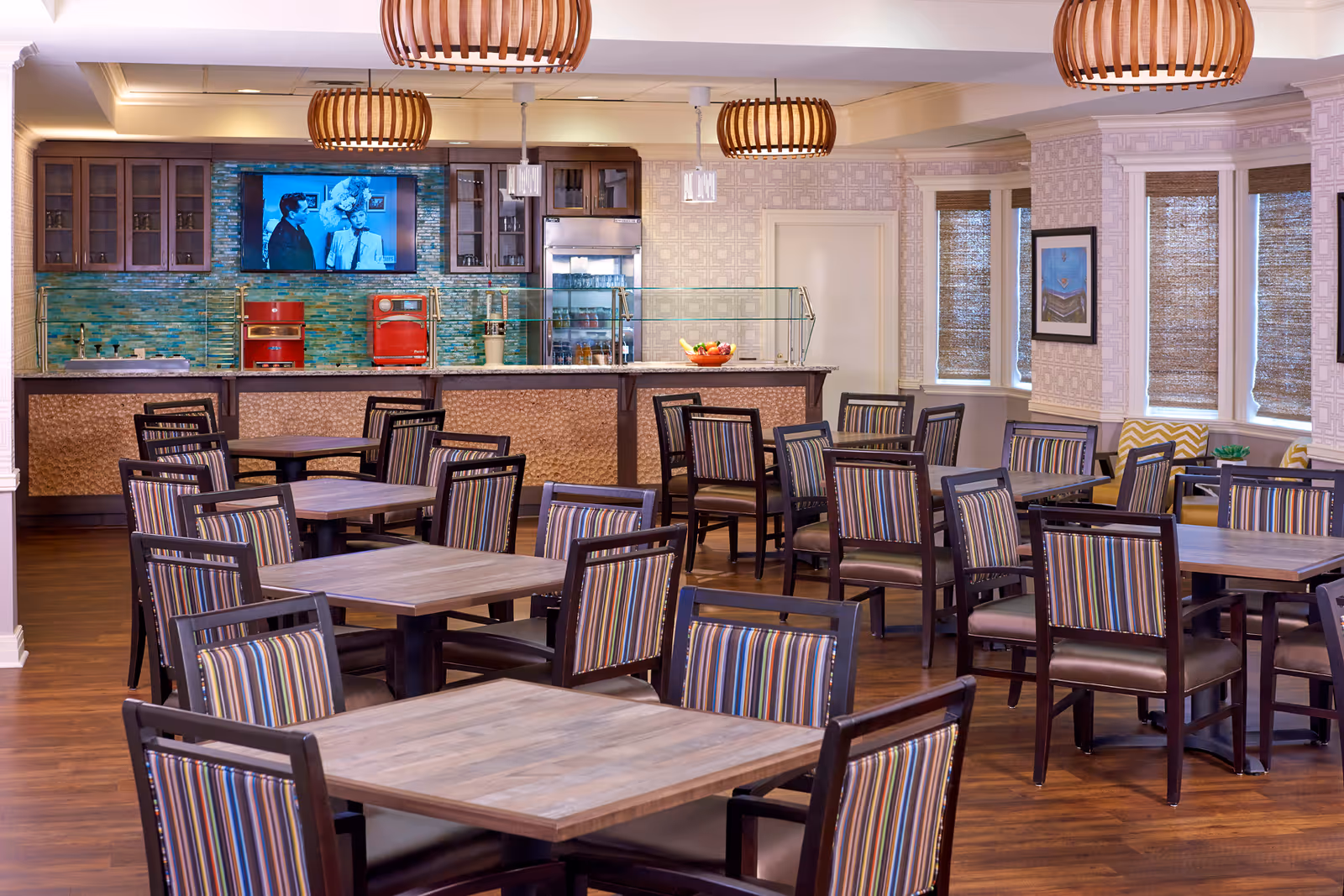 A dining room in an assisted living facility featuring multiple wooden tables with striped cushioned chairs. The room has a serving counter with a glass sneeze guard, red kitchen appliances, a refrigerator, and a wall-mounted television playing a black and white show. The space is warmly lit with wooden pendant lights and has windows with woven shades and framed artwork on the walls.