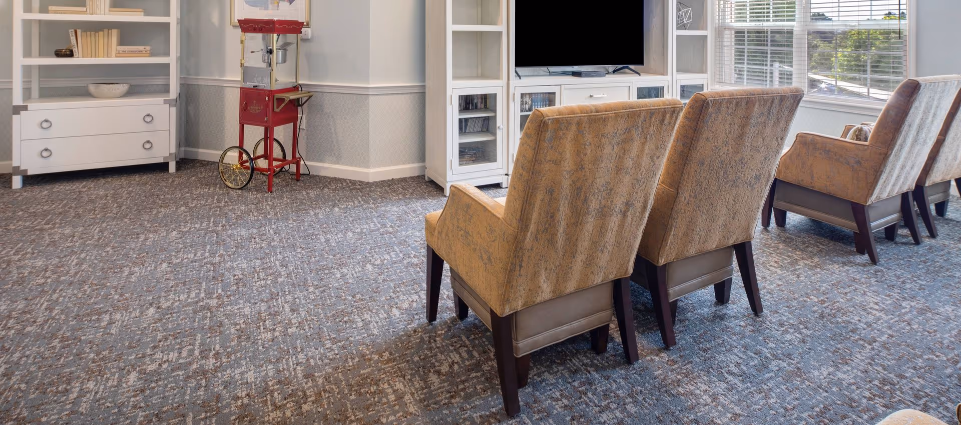 A cozy living room area with four upholstered armchairs arranged in two rows facing a white entertainment center with a flat-screen TV. To the left, there is a white bookshelf with books and decorative items, and a vintage-style red popcorn machine on wheels. Large windows with blinds allow natural light to brighten the room.