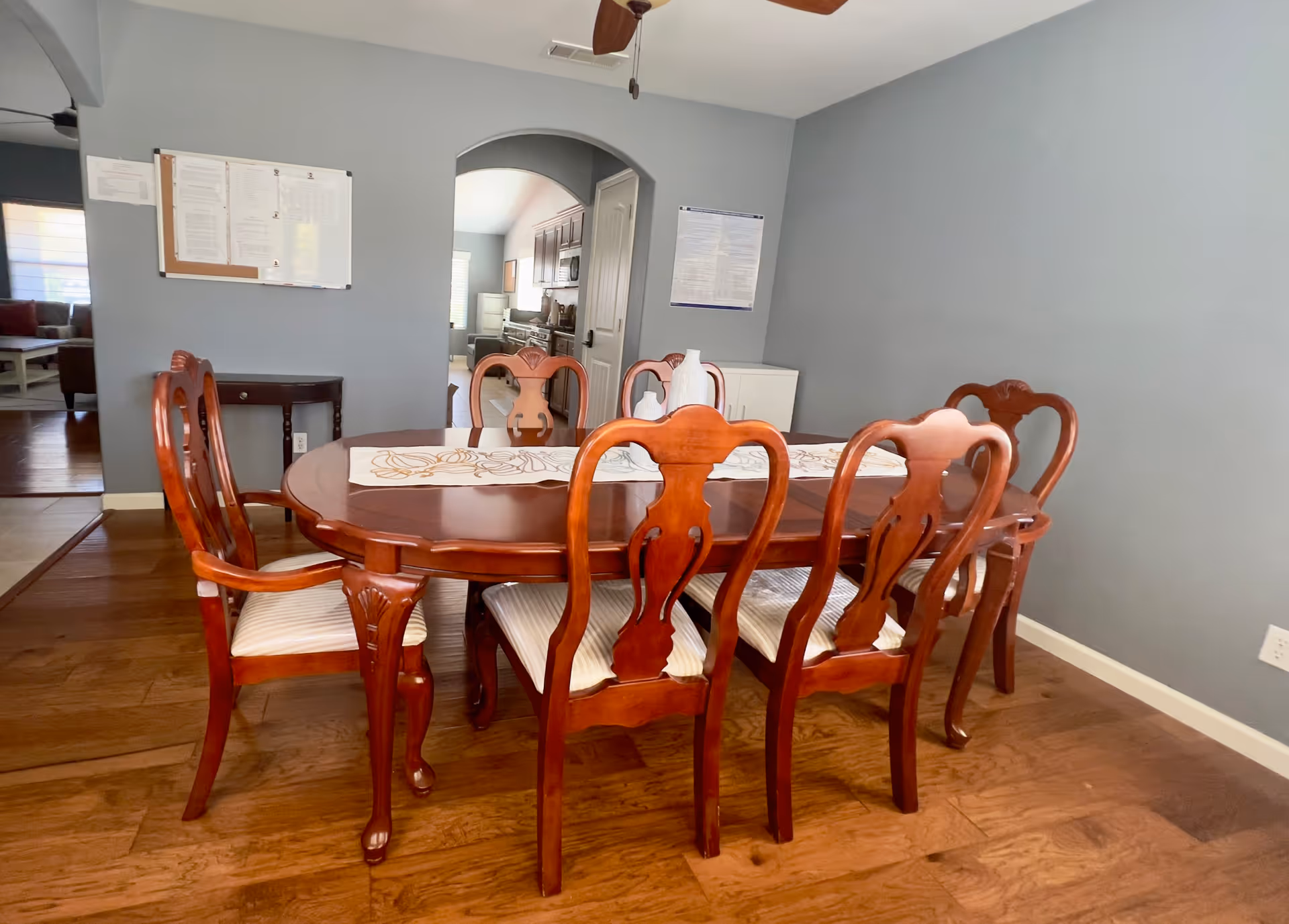 A dining room with a wooden dining table and six matching chairs with cushioned seats. The table has a decorative runner and a white vase on it. The room has wooden flooring and light blue walls. In the background, there is an archway leading to a kitchen area with cabinets and appliances visible.
