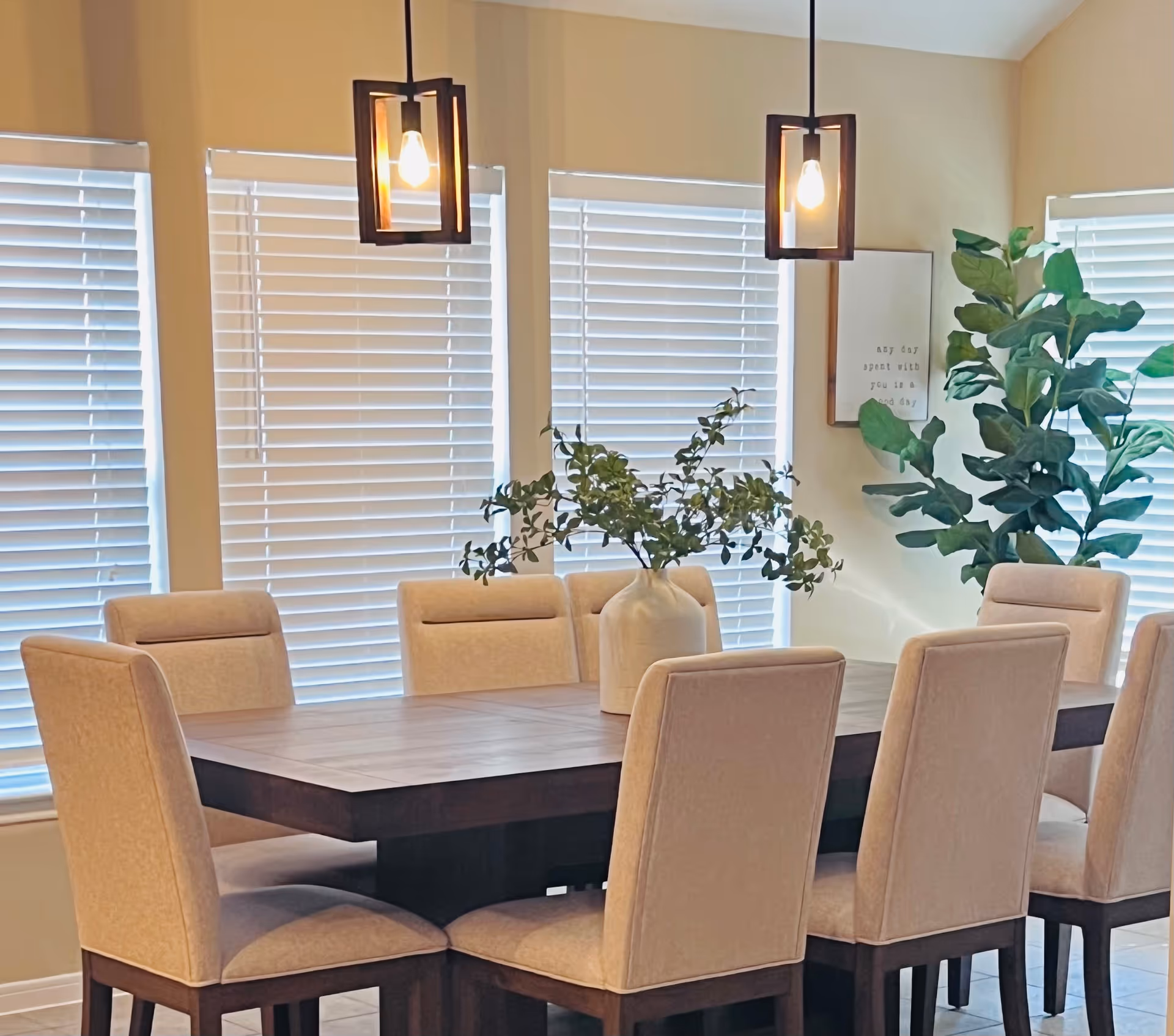 A dining room with a large rectangular wooden table surrounded by eight beige upholstered chairs. Two modern pendant lights hang above the table. A large vase with green leafy branches is placed in the center of the table. The room has several windows with white blinds and a large green plant in the corner. A framed sign with text is visible on the wall.