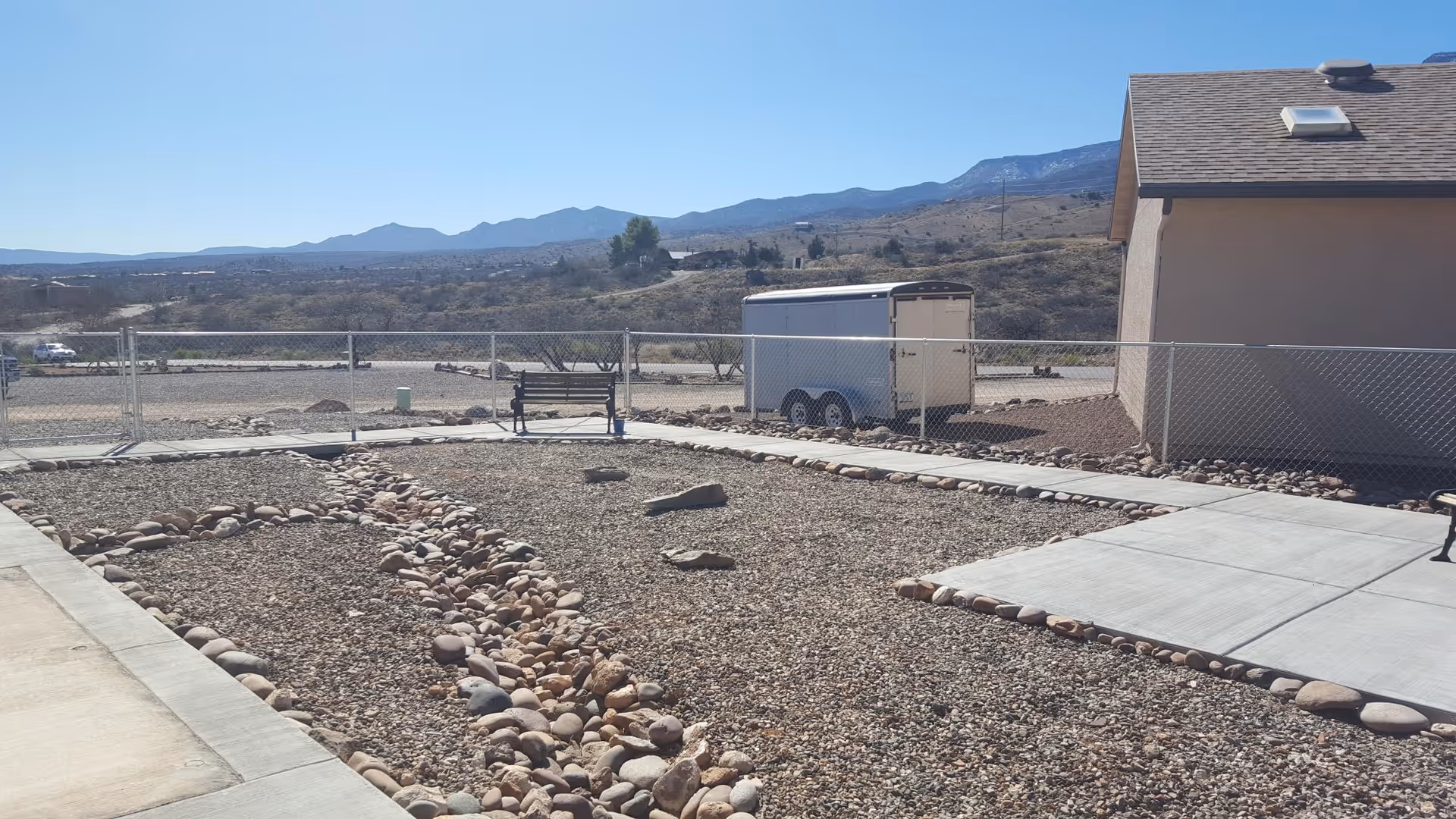 Outdoor area with a gravel ground and a pathway bordered by rocks. There is a metal bench near a chain-link fence, and a white trailer is parked behind the fence. In the background, there are hills and mountains under a clear blue sky.