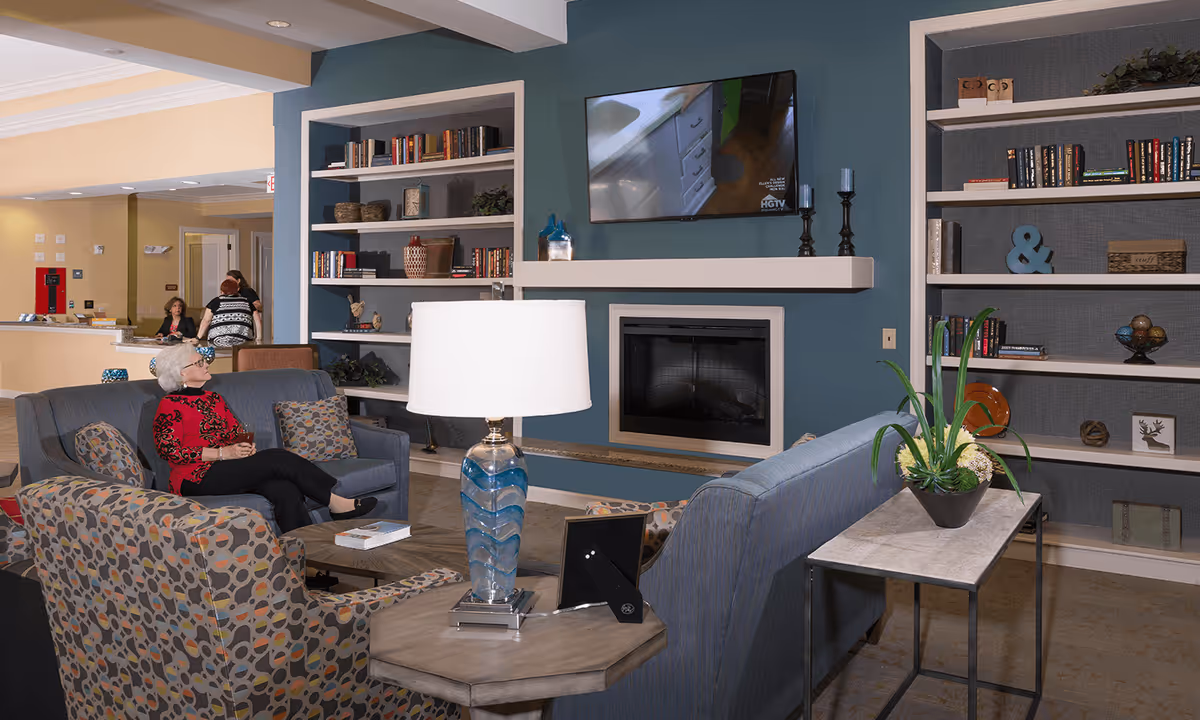 A cozy living room area in a senior living facility with a woman sitting on a blue couch. The room features built-in bookshelves filled with books and decorative items, a wall-mounted TV above a fireplace, a patterned armchair, a side table with a blue glass lamp, and a small table with a potted plant. In the background, two women are seen near a reception desk.