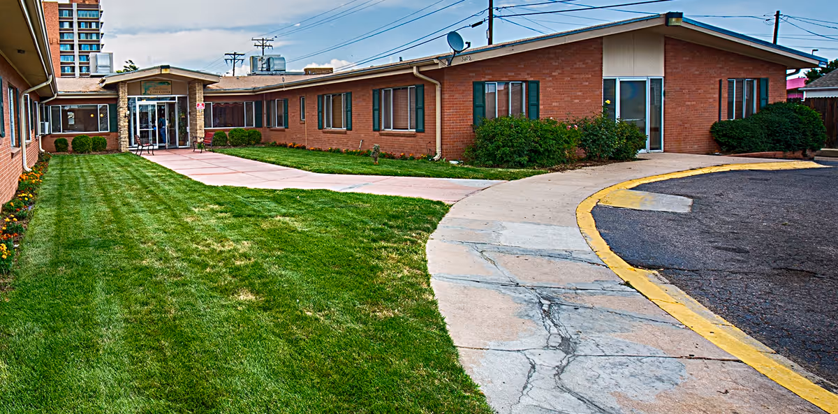 Exterior view of a single-story brick building with a well-maintained lawn and a curved sidewalk leading to the entrance of Parkview Care Center. The building has multiple windows and some bushes near the walls. The sky is partly cloudy.