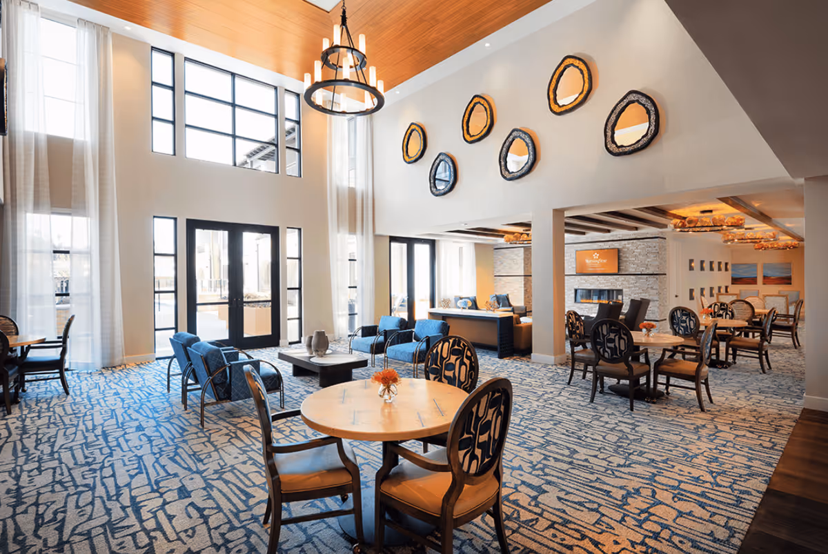 Bright two-story community lounge and dining area with round tables, seating groups, large windows, and decorative wall art.