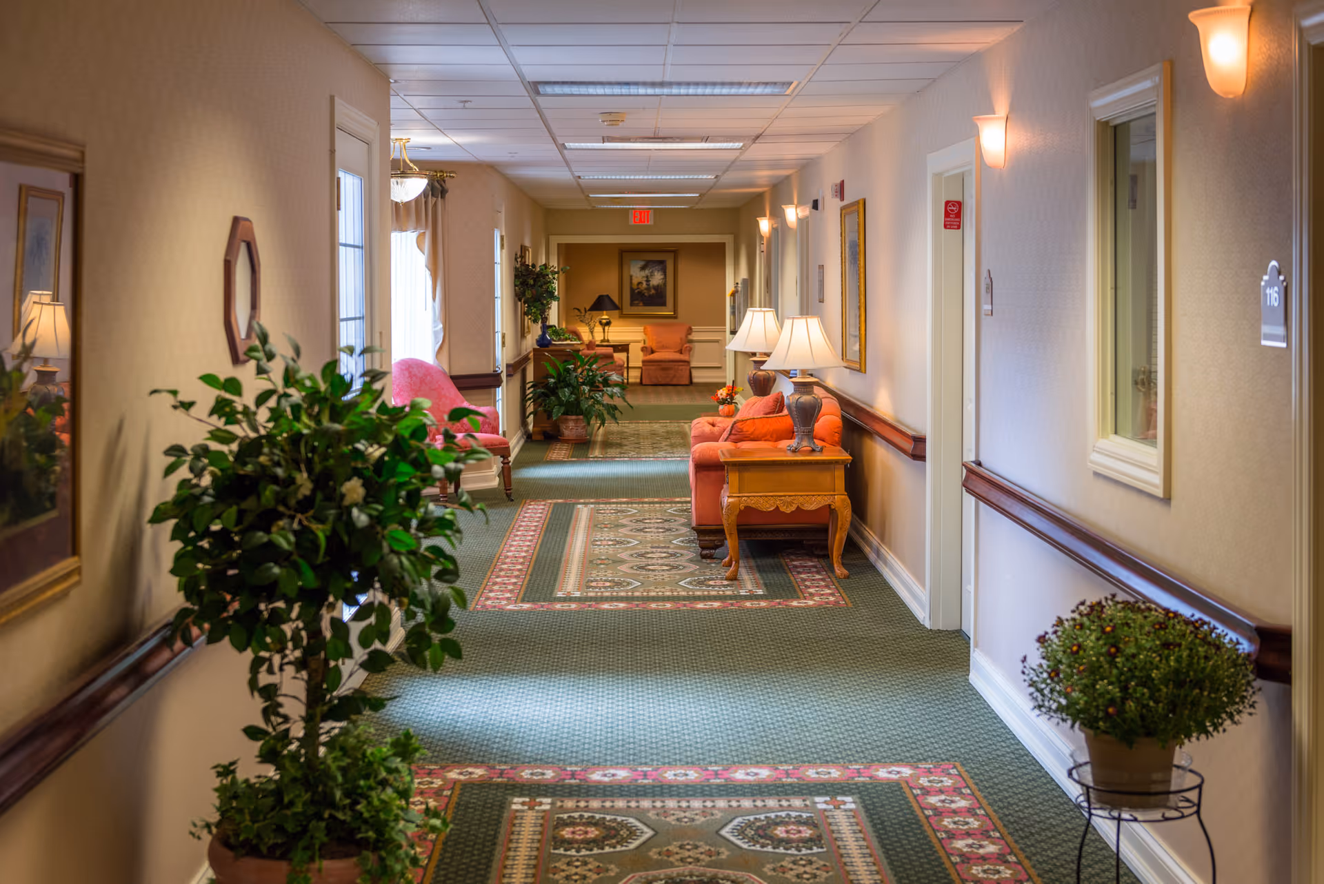 A well-lit hallway in a senior living facility with patterned green carpet and beige walls. The hallway is decorated with potted plants, framed artwork, wall sconces, and several seating areas featuring orange upholstered chairs and wooden side tables with lamps. Doors to rooms line the right side of the hallway.