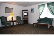 A living room with two green upholstered armchairs and a matching green sofa. There is a wooden coffee table in front of the sofa and a side table with a lamp between the armchairs. A small TV is placed on a wooden stand in the corner, with a few books on the shelves below. The room has carpeted flooring, white walls, a framed picture, and a window with green lace curtains.