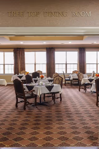 Interior view of the Tree Top Dining Room with multiple tables covered in white tablecloths and black table runners, set with dining ware and small floral centerpieces. The room has large windows with brown curtains allowing natural light to fill the space, and patterned carpet flooring.