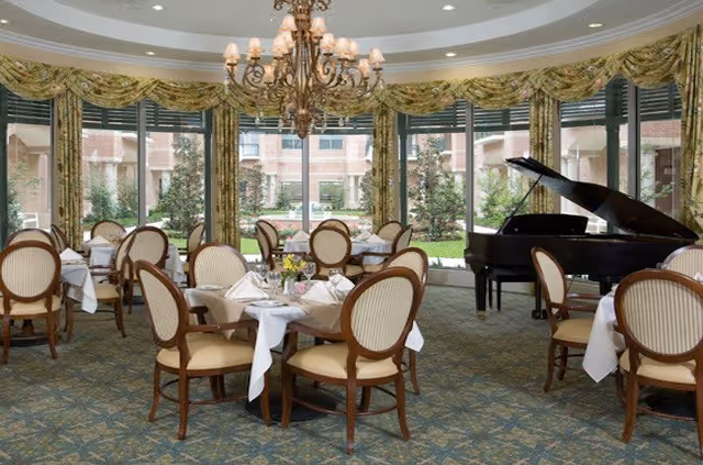Elegant dining room with round tables covered in white tablecloths and set with napkins and glassware. The room features large windows with floral curtains overlooking a garden, a grand chandelier hanging from the ceiling, and a black grand piano in the corner.