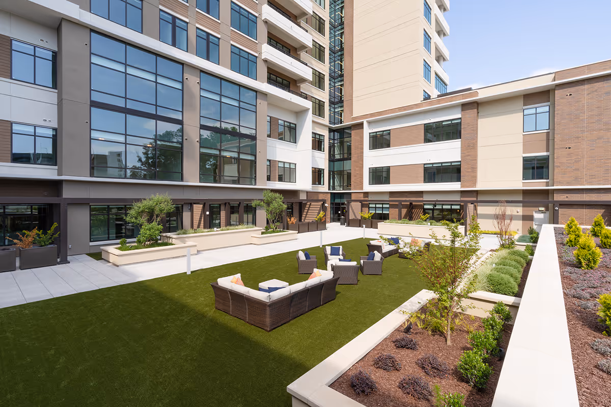 Outdoor courtyard area at The Cardinal at North Hills featuring green artificial turf, several wicker seating arrangements with cushions, surrounded by landscaped garden beds and modern multi-story building with large windows in the background.