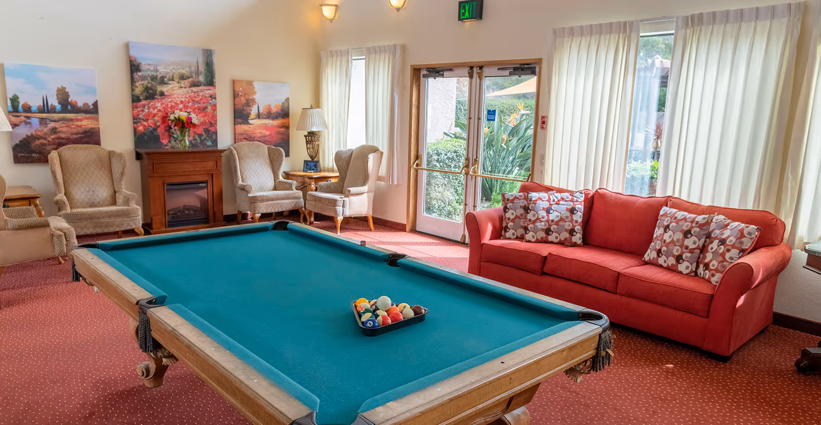 Bright common room with a pool table in the center, a red sofa, armchairs, artwork, and glass doors/windows.