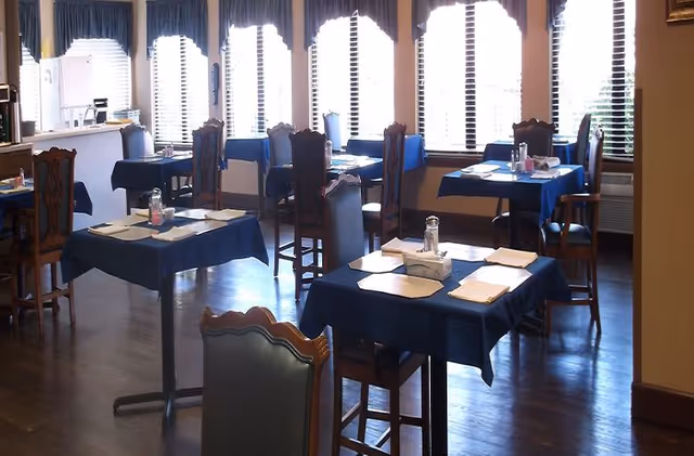 Dining room with multiple tables covered in blue tablecloths, each set with napkins, salt and pepper shakers, and tissue boxes. Wooden chairs surround the tables, and large windows with blinds allow natural light to fill the room.