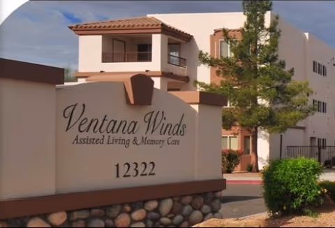 Exterior view of Ventana Winds Assisted Living & Memory Care facility with a sign displaying the name and address 12322, a multi-story building, trees, and a clear sky.