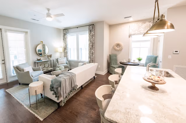 Bright open-concept living area with a sofa and armchairs, a coffee table, adjacent kitchen island with bar stools, and a small dining nook.