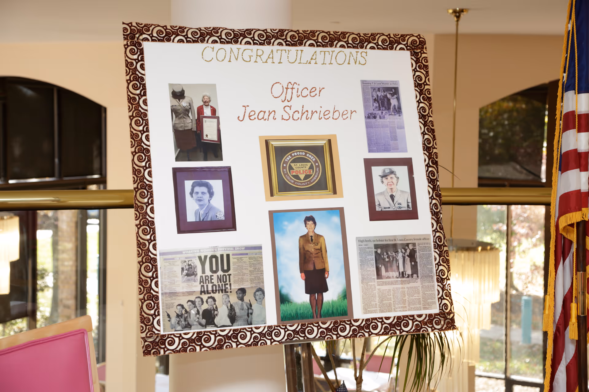 A congratulatory display board for Officer Jean Schrieber featuring multiple photographs, newspaper clippings, and a police badge emblem. The board is decorated with a patterned border and is set up indoors near windows and an American flag.