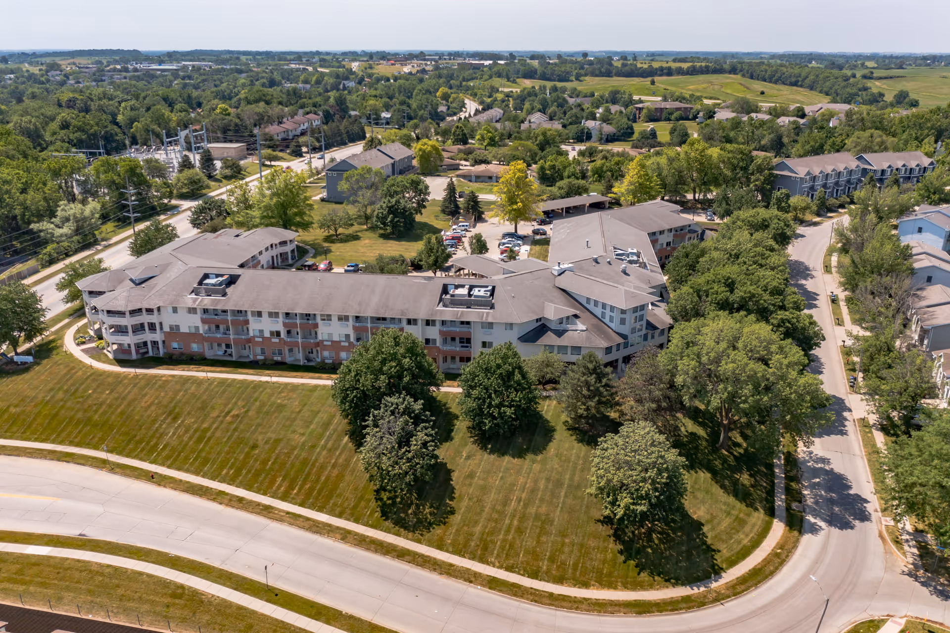 Aerial view of a multi-story senior living building surrounded by lawns, trees, parking areas and curved roads.