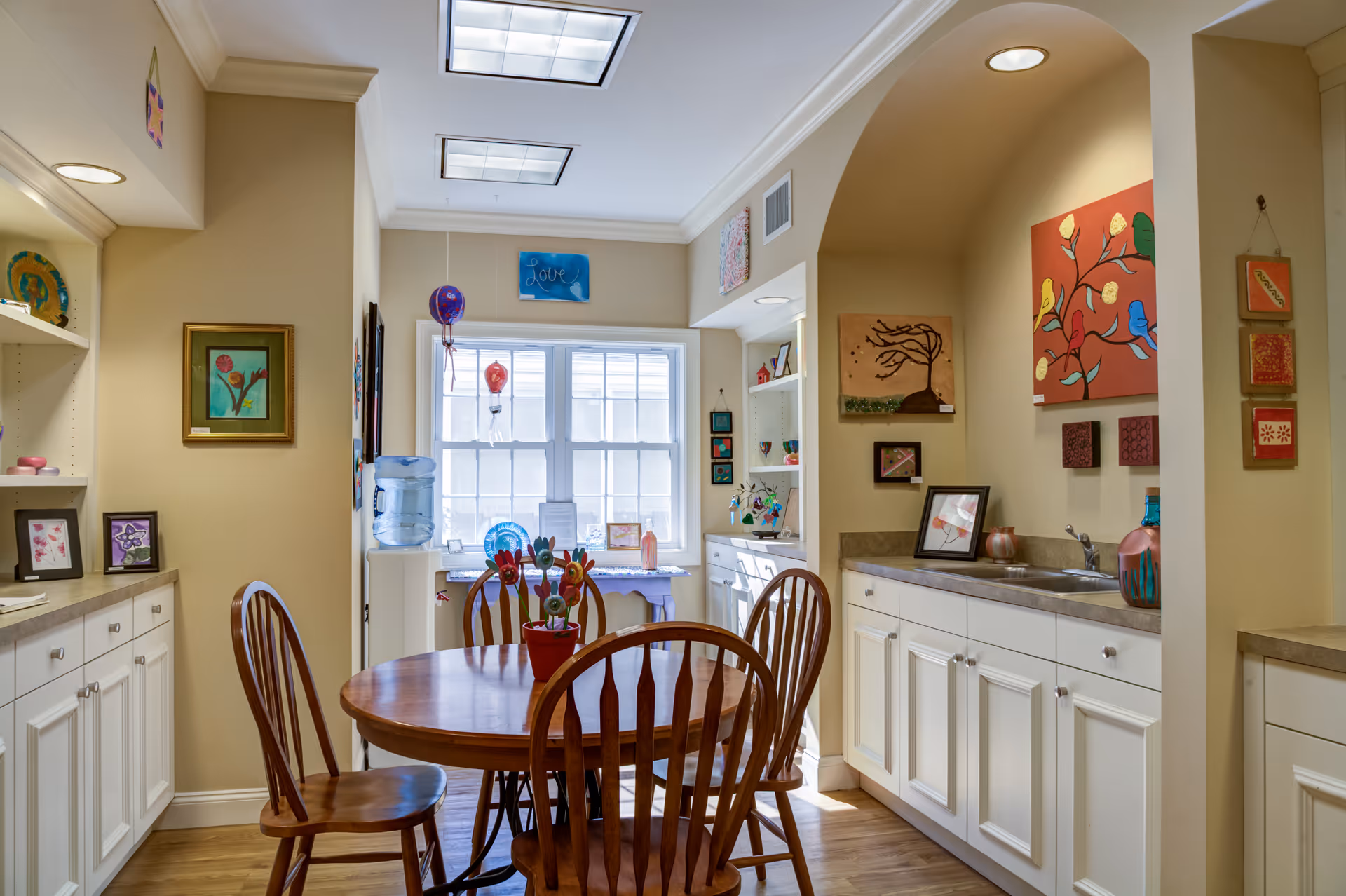 A cozy dining area with a round wooden table and four wooden chairs. The room features white cabinetry along the walls, colorful artwork, and decorative items on the countertops and shelves. A window at the far end lets in natural light, and a water cooler is positioned near the window.