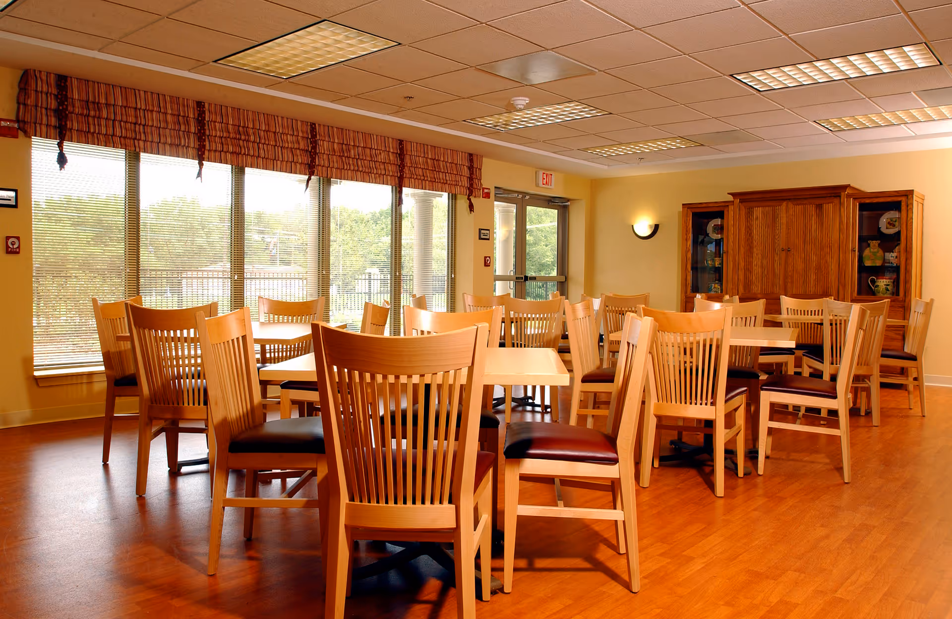A bright dining room with multiple wooden tables and chairs arranged neatly. Large windows with blinds and valances allow natural light to fill the room. A wooden cabinet with glass doors is visible against the far wall, and the floor is covered with wood-like flooring.