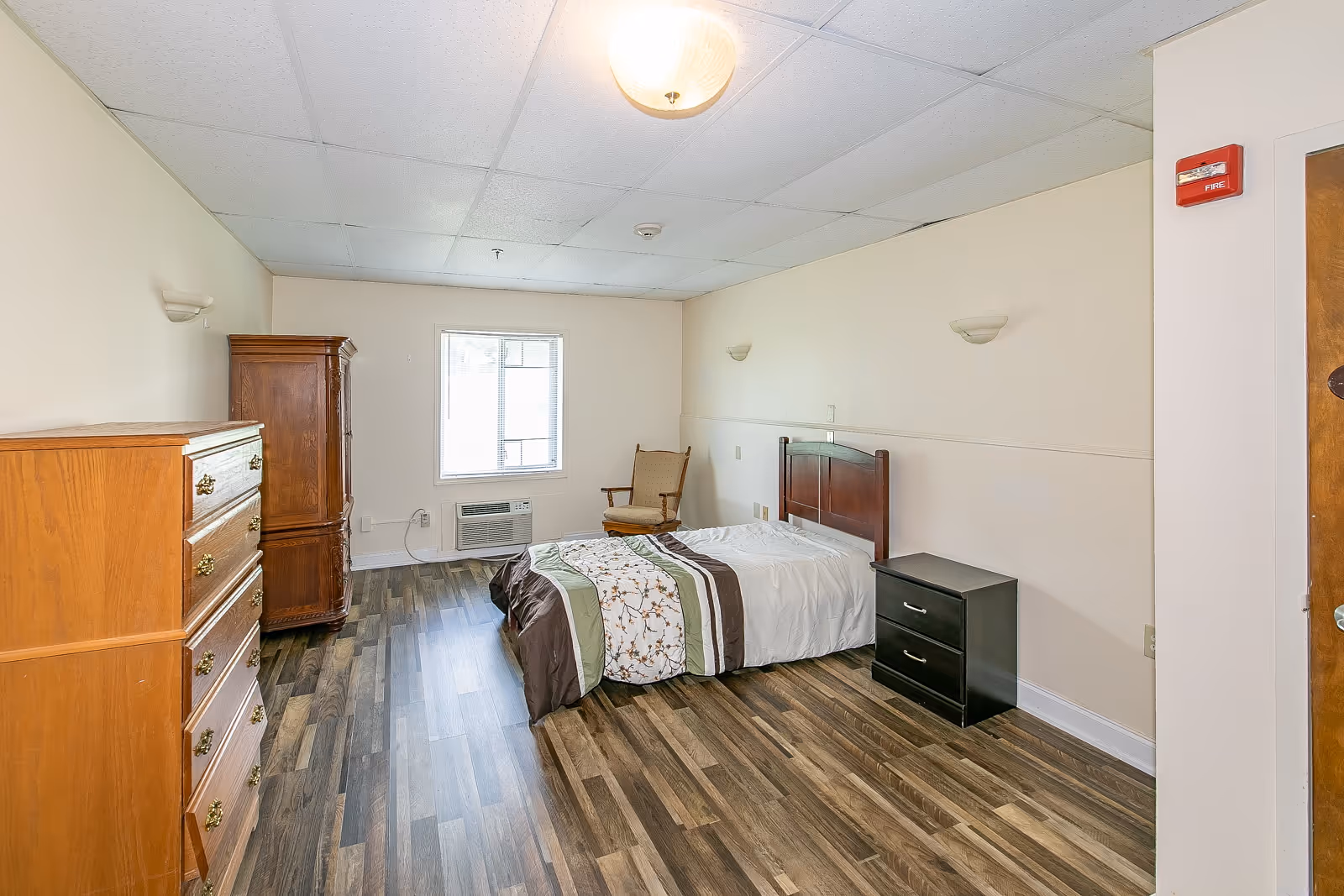 A simple senior living bedroom with a single bed covered with a white and brown patterned comforter, a wooden headboard, a black nightstand with two drawers, a wooden dresser, a wooden armoire, a wooden chair, and a window with an air conditioning unit below it. The room has light-colored walls and wood-patterned flooring.