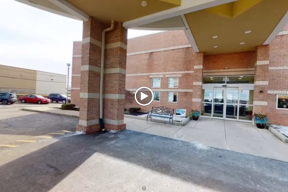 Covered drop-off entrance to a brick assisted living building with glass double doors, a bench, and potted plants.
