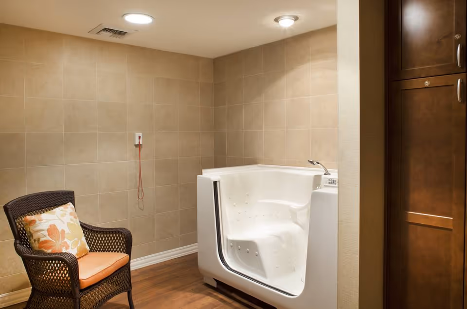 Tiled room with a white walk-in bathtub, a wicker chair with a floral cushion, and an emergency pull cord on the wall.