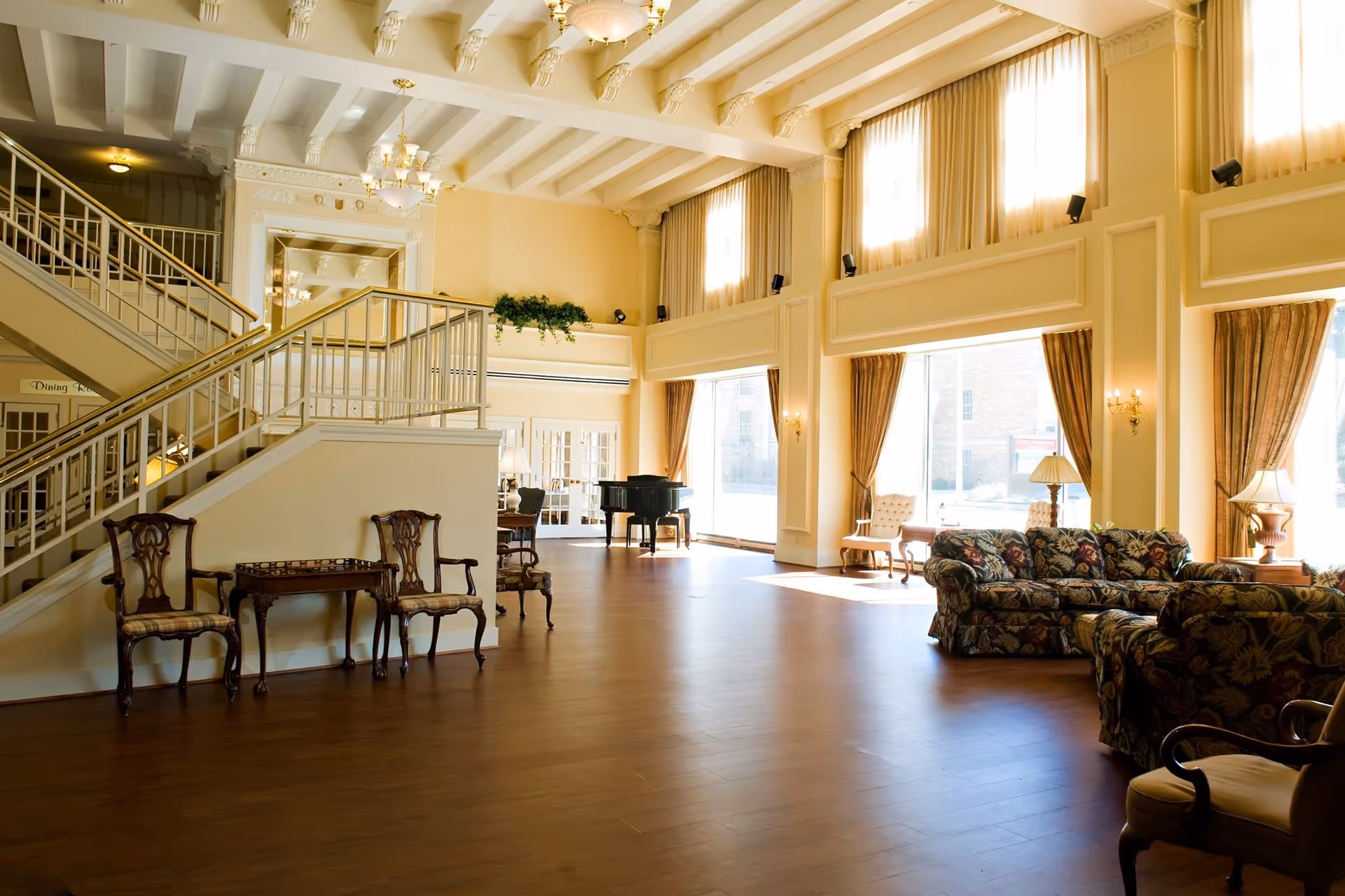 A spacious, well-lit senior living community common area with high ceilings, large windows with curtains, wooden flooring, and classic furniture including floral-patterned sofas, wooden chairs, a small table, and a black grand piano near the back. A staircase with white railings is visible on the left side.