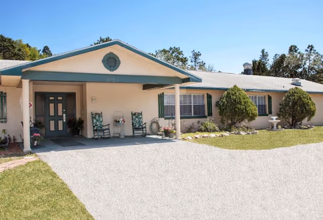 Front exterior of a single-story senior living building with a covered entrance, rocking chairs, and landscaped lawn.
