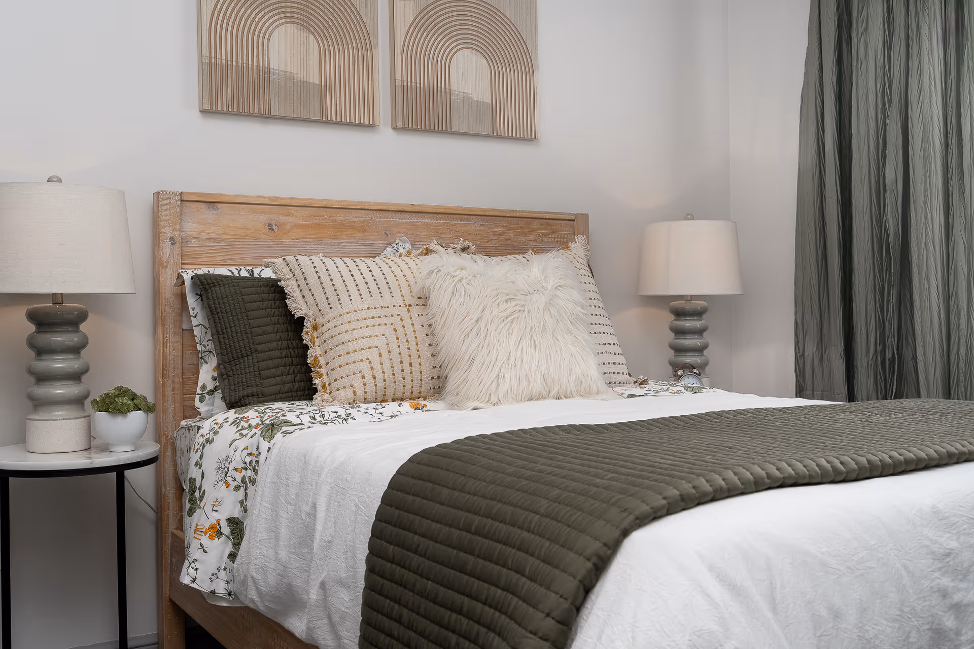 A neatly made bed with a wooden headboard, adorned with multiple decorative pillows including a fluffy white one and patterned ones. The bed has a white sheet with a floral pattern and a dark green quilted blanket. On each side of the bed are matching bedside tables with gray ceramic lamps and a small plant on the left table. Two abstract wooden wall art pieces hang above the headboard, and dark green curtains are visible on the right side.