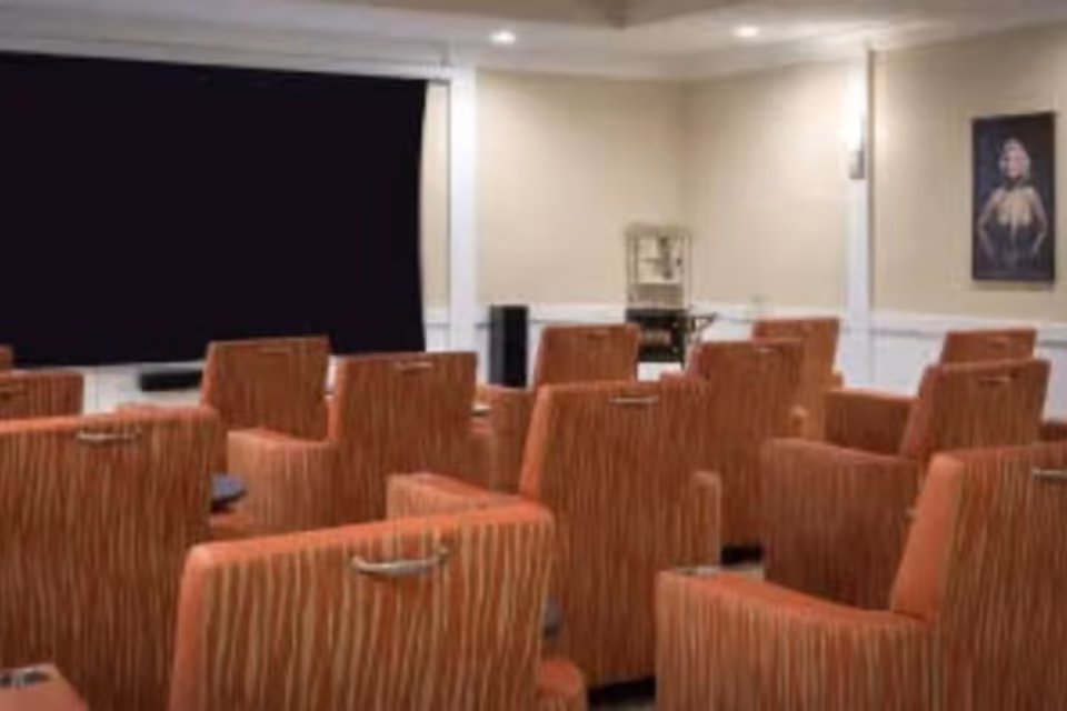 A small theater room with multiple rows of orange patterned armchairs facing a large blank black screen on the wall. The room has beige walls with white trim, recessed ceiling lights, and a framed portrait on the right wall.