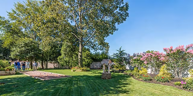 A sunny outdoor garden area with a well-maintained lawn, a large tree, a small wishing well structure, and a bocce ball court. Several people are gathered near the bocce ball court. The garden is bordered by a wooden fence and flowering bushes with pink blossoms.